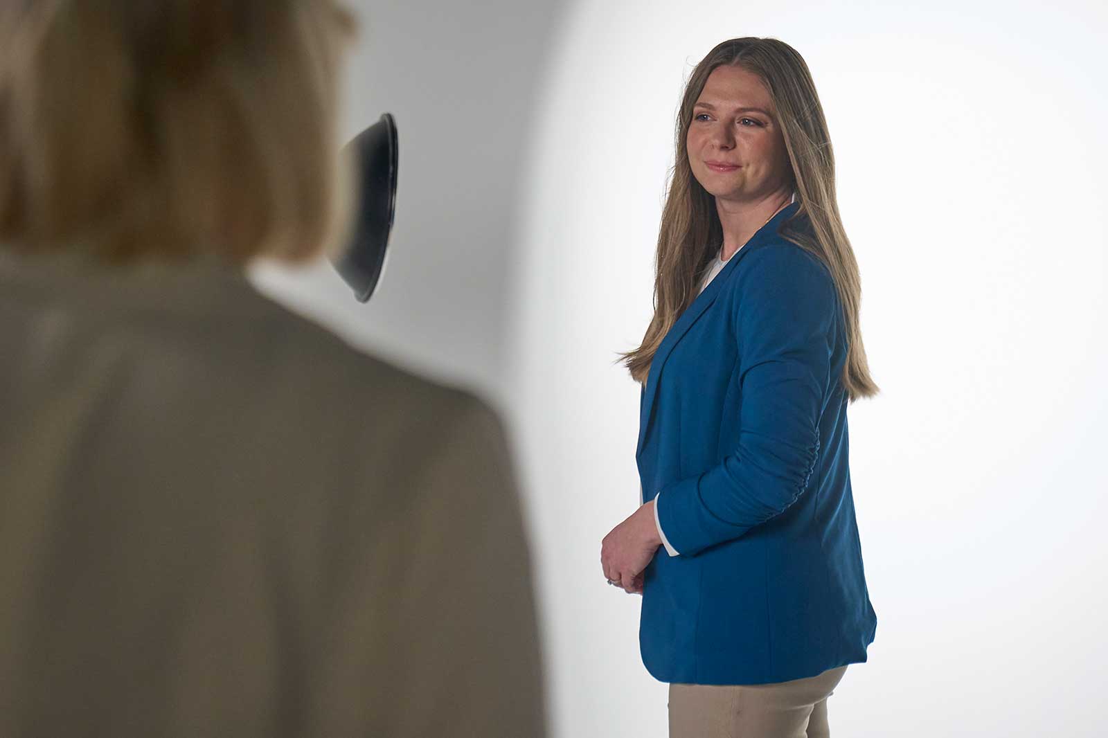 A person with long brown hair wears a blue jacket and stands against a light background.
